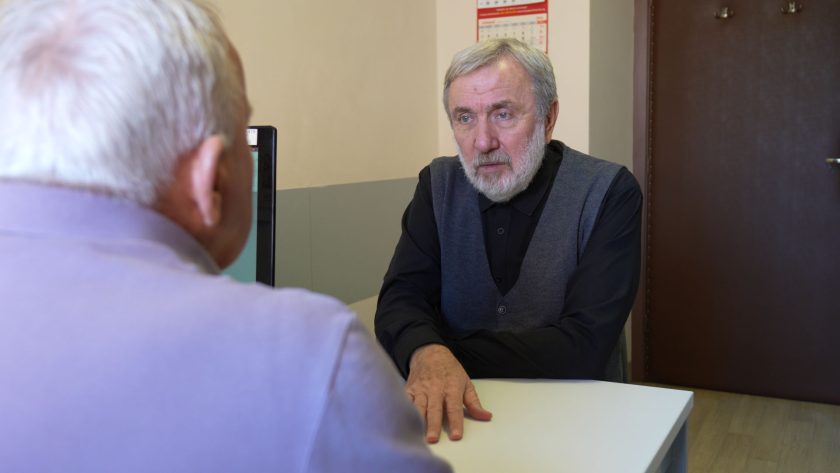 Lekarz w ciemnym ubraniu rozmawia z pacjentem w gabinecie. Starszy mężczyzna w fioletowej koszulce siedzi przy biurku, a jego rozmówca uważnie słucha, patrząc mu w oczy