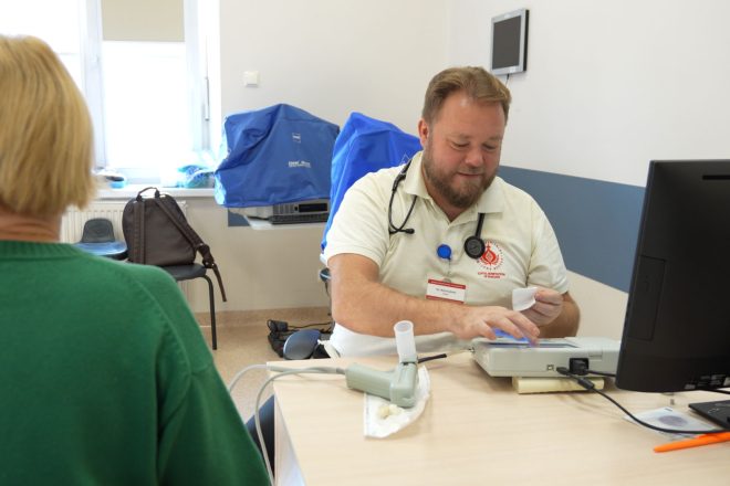 Lekarz wykonuje badanie spirometryczne u pacjentki. Mężczyzna w białym stroju analizuje wyniki na urządzeniu, a kobieta siedzi tyłem do obiektywu. W tle widoczne są okno i sprzęt medyczny