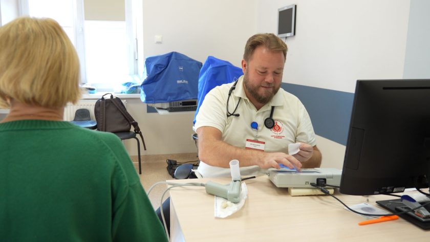 Lekarz wykonuje badanie spirometryczne u pacjentki. Mężczyzna w białym stroju analizuje wyniki na urządzeniu, a kobieta siedzi tyłem do obiektywu. W tle widoczne są okno i sprzęt medyczny