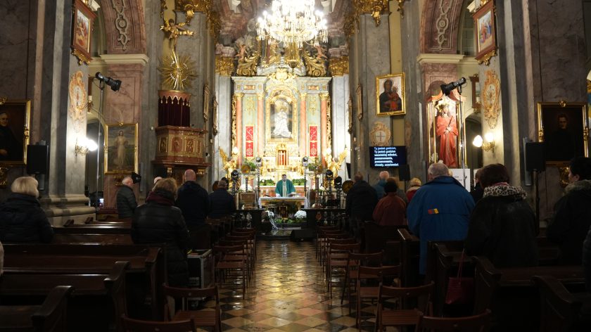 Ołtarz kościoła ozdobiony kwiatami i świecami, ksiądz celebruje mszę. W tle monumentalna dekoracja ołtarza i rozświetlony żyrandol