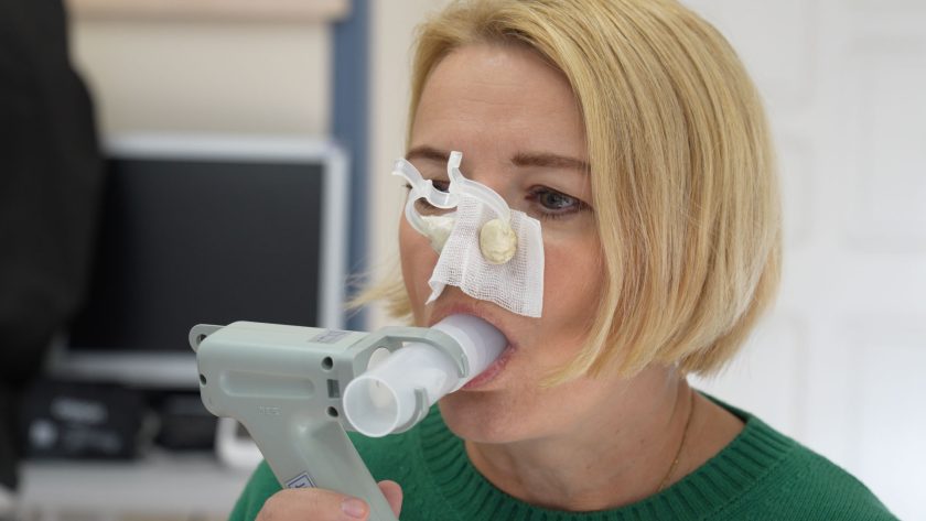 Kobieta w zielonym swetrze wykonuje badanie spirometryczne. Trzyma ustnik urządzenia spirometrycznego w ustach, a na nosie ma założone klipsy uszczelniające. W tle rozmyte elementy wyposażenia gabinetu