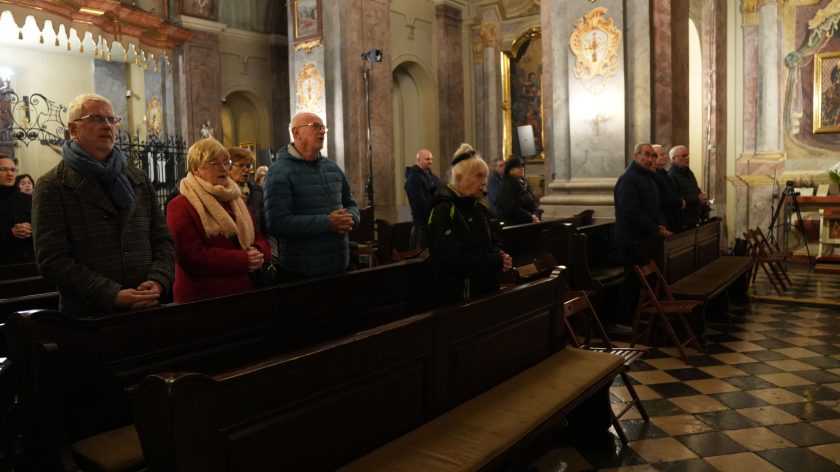 Ławki wypełnione wiernymi w różnym wieku. Na pierwszym planie kobieta w czerwonym płaszczu i mężczyzna w okularach oraz szalu