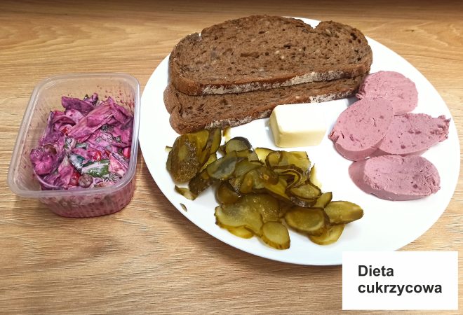 Na zdjęciu przedstawiono posiłek z kategorii "Dieta cukrzycowa" podany na białym talerzu, również na jasnym, drewnianym blacie. Na talerzu znajdują się ciemne kromki chleba żytniego oraz kawałek masła, które można użyć jako dodatek. Po prawej stronie chleba leżą plastry pasztetu drobiowego w jasnoróżowym kolorze, który jest miękki i delikatny. Po lewej stronie talerza widzimy porcję ogórków konserwowych w cienkich plasterkach. Obok talerza, w plastikowym pojemniku, znajduje się kolorowa sałatka z czerwonej kapusty, z dodatkiem papryki i warzyw zielonych, wszystko połączone jasnym sosem. W prawym dolnym rogu zdjęcia widnieje napis "Dieta cukrzycowa"