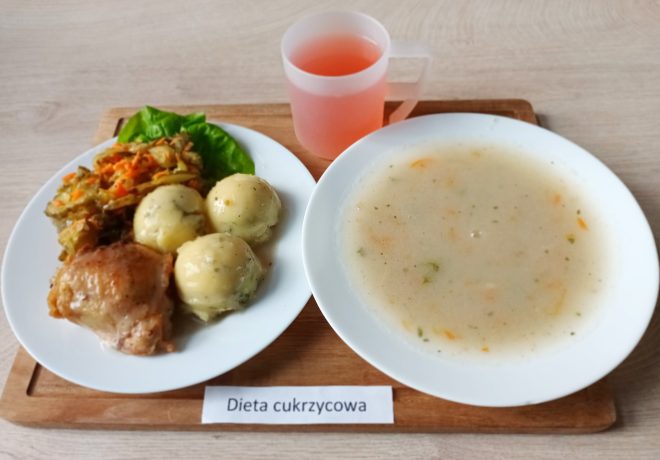 Zdjęcie przedstawia zestaw obiadowy oznaczony etykietą "Dieta cukrzycowa" na drewnianej tacce. Po lewej stronie na białym talerzu znajduje się pieczone udko z kurczaka, trzy kluseczki ziemniaczane posypane ziołami oraz porcja mieszanki warzywnej, składającej się z marchewki, kapusty i innych warzyw, ułożonej na liściu sałaty. Obok, po prawej stronie, znajduje się głęboki talerz z zupą o jasnym bulionie z widocznymi kawałkami warzyw, takimi jak marchewka i natka pietruszki. W górnej części tacy stoi przezroczysty kubek z napojem o różowym odcieniu