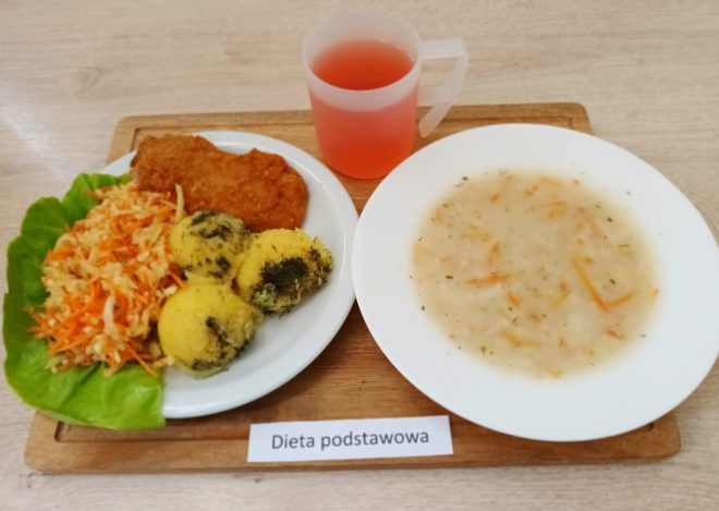 Posiłek obiadowy ustawiony na drewnianej desce, z napisem „Dieta podstawowa”. Po lewej stronie talerza znajduje się panierowany kotlet, porcja surówki z marchwi i kapusty na liściu sałaty oraz trzy kulki puree ziemniaczanego posypane koperkiem. Po prawej stronie umieszczono talerz z zupą, prawdopodobnie jarzynową, o kremowej konsystencji z widocznymi kawałkami marchewki i przyprawami. Obok, w przezroczystym kubku, znajduje się czerwony napój