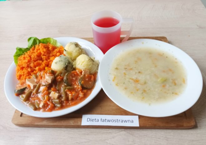 Na zdjęciu znajduje się zestaw obiadowy przeznaczony dla osoby na diecie łatwostrawnej. Na dużym białym talerzu po lewej stronie znajdują się ziemniaki puree w trzech kulkach, obok podana jest mieszanka warzyw duszonych w sosie pomidorowym z kawałkami mięsa, fasolką szparagową i plasterkami ogórków. Danie uzupełnia porcja ugotowanej marchewki, ułożona na liściu sałaty. Obok, na oddzielnym talerzu, znajduje się zupa warzywna o jasnym wywarze z kawałkami marchwi i innych warzyw. W zestawie znajduje się również przezroczysty kubek z czerwoną herbatą lub kompotem. Na drewnianej desce, na której stoi zestaw, widnieje napis „Dieta łatwostrawna”