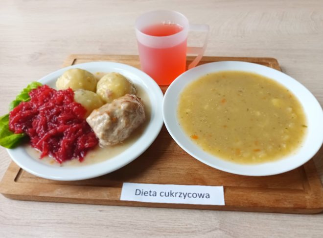 Na drewnianej tacy widoczny jest posiłek oznaczony jako „Dieta cukrzycowa”. Po lewej stronie tacy znajduje się talerz z głównym daniem: trzy ziemniaki w mundurkach, porcja buraków na ciepło oraz kawałek mięsa w jasnym sosie. Po prawej stronie znajduje się miseczka z gęstą, żółtawą zupą jarzynową, w której widoczne są kawałki warzyw. U góry tacy stoi przezroczysty kubek z czerwonym napojem, prawdopodobnie kompotem