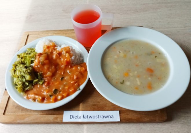 Na drewnianej desce znajduje się zestaw obiadowy opisany jako "Dieta łatwostrawna". Z lewej strony talerz z porcją gotowanego ryżu, brokułami oraz sosem warzywnym o pomidorowej bazie, z widocznymi kawałkami warzyw, który polewa ryż. Obok, po prawej stronie, głęboki talerz z zupą o kremowej konsystencji z drobno pokrojonymi warzywami, takimi jak marchew, ziemniaki i pietruszka. Na górze zdjęcia przezroczysty kubek z różowym napojem