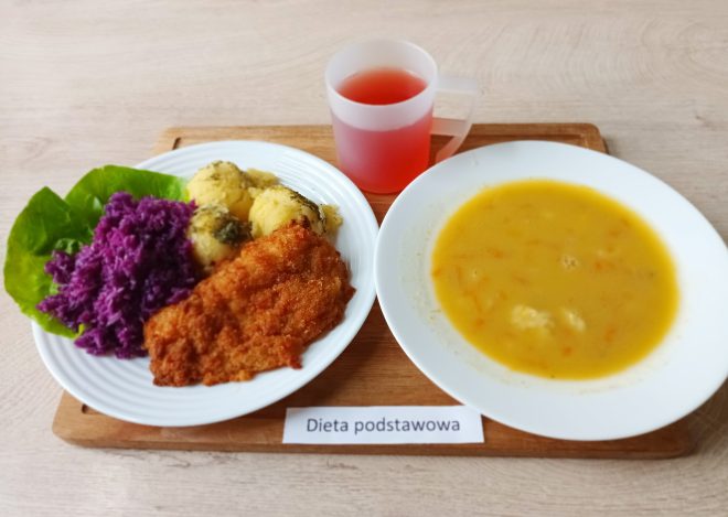 Zdjęcie przedstawia obiad w wersji „Dieta Podstawowa”. Na białym talerzu znajduje się kotlet panierowany, porcja purée ziemniaczanego posypana koperkiem, a także surówka z czerwonej kapusty oraz liść sałaty dla dekoracji. Obok, w oddzielnym głębokim talerzu, serwowana jest zupa jarzynowa o intensywnie żółtym kolorze, z widocznymi kawałkami marchewki i warzyw. Do posiłku dołączony jest czerwony napój w przezroczystym kubku, który uzupełnia ten klasyczny zestaw obiadowy