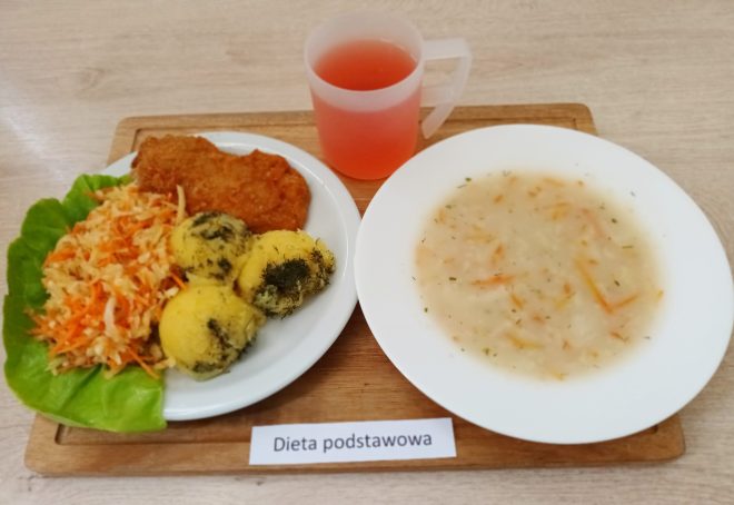 Na drewnianej desce leżą dwie białe zastawy z obiadem oraz kubek z czerwonawym napojem. Na pierwszym talerzu: panierowany kotlet, puree ziemniaczane posypane koperkiem, surówka z marchewki i kapusty na liściu sałaty. Na drugim talerzu widoczna jest miseczka z lekką zupą jarzynową. Na desce znajduje się etykieta z napisem "Dieta podstawowa"