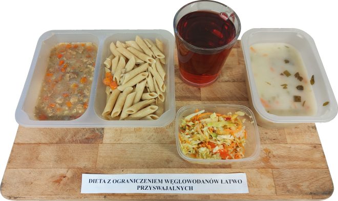 Na drewnianej desce podany jest zestaw obiadowy w pojemnikach. W jednym znajduje się porcja gulaszu z warzywami, w drugim pełnoziarnisty makaron penne z kawałkami marchewki, a w kolejnym - kremowa zupa jarzynowa z ziołami. Obok znajduje się mały pojemnik z surówką z białej kapusty, marchwi i koperku. W tle szklanka z czerwonym napojem. Na desce widnieje napis "Dieta z ograniczeniem węglowodanów łatwo przyswajalnych"