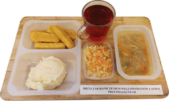 Dieta z ograniczeniem węglowodanów łatwo przyswajalnych: zestaw obiadowy na drewnianej tacy. Na pierwszym planie leżą panierowane paluszki rybne oraz porcje puree ziemniaczanego. Obok, w przezroczystym pojemniku, surówka z kapusty z dodatkiem marchwi. Zestaw uzupełnia przezroczysty kubek z czerwonym napojem oraz zupa jarzynowa w plastikowym pojemniku. Na tacy widoczna etykieta z napisem „Dieta z ograniczeniem węglowodanów łatwo przyswajalnych”