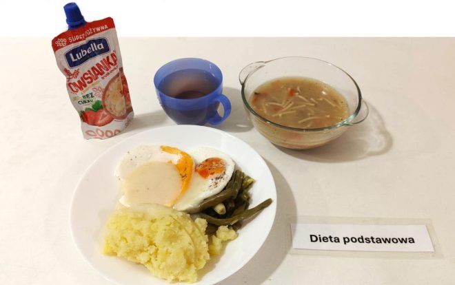 Na zdjęciu widzimy obiad oznaczony jako „Dieta podstawowa”. Na białym talerzu znajduje się porcja purée ziemniaczanego, dwa jajka sadzone oraz zielona fasolka szparagowa. Obok, w niebieskim kubku, znajduje się ciemny napój, najprawdopodobniej herbata. W przezroczystej miseczce po prawej stronie serwowana jest zupa z kawałkami marchewki i cienkim makaronem. Obok posiłku znajduje się saszetka owsianki owocowej Lubella, prawdopodobnie do samodzielnego przygotowania