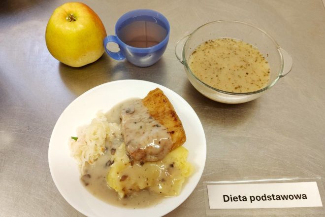 Dieta podstawowa: obiad na stalowym blacie. Na talerzu znajdują się panierowana pierś z kurczaka w sosie pieczarkowym, puree ziemniaczane oraz kapusta kiszona. Obok, w szklanej miseczce, widoczna porcja żurku. Zestaw uzupełniają jabłko oraz napój w niebieskim kubku. Na blacie umieszczona jest etykieta z napisem „Dieta podstawowa”