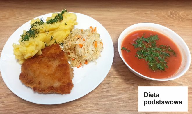 Na zdjęciu zaprezentowano obiad w ramach diety podstawowej. Na białym talerzu znajdują się: panierowany kotlet, porcja puree ziemniaczanego z posypką z koperku oraz porcja surówki z białej kapusty z dodatkiem marchewki. Obok talerza umieszczono miskę z zupą pomidorową, ozdobioną świeżymi listkami natki pietruszki. W prawym dolnym rogu widnieje etykieta z napisem „Dieta podstawowa”