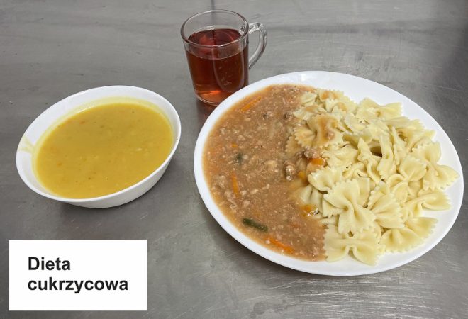 Na stole widoczny jest zestaw obiadowy. Na głównym talerzu znajduje się porcja ugotowanego makaronu kokardki, obok gulasz z mielonym mięsem i warzywami, podany w aromatycznym sosie. Obok talerza stoi miseczka z kremową zupą jarzynową w żółtym odcieniu oraz szklanka z herbatą. W lewym dolnym rogu zdjęcia znajduje się napis "Dieta cukrzycowa"