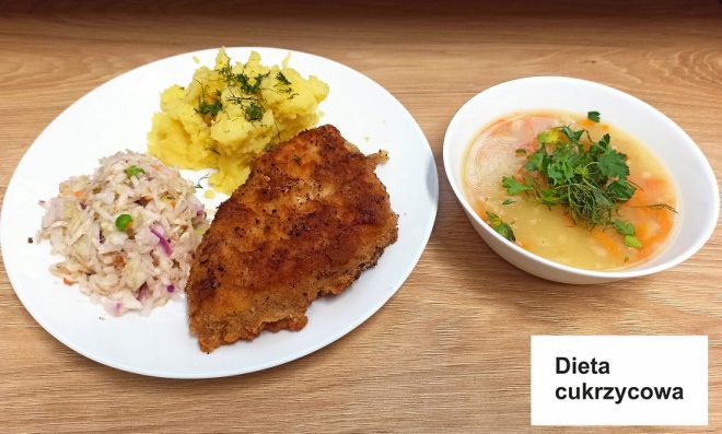 Dieta cukrzycowa Zdjęcie przedstawia pełnowartościowy obiad oznaczony jako "Dieta cukrzycowa". Na białym talerzu znajduje się panierowany filet drobiowy, puree ziemniaczane posypane świeżym koperkiem oraz surówka z białej kapusty z dodatkiem marchewki, groszku i cebuli. Obok, w białej miseczce, podano lekką zupę jarzynową z widocznymi kawałkami marchewki i ziemniaków, udekorowaną natką pietruszki. Całość przeznaczona jest dla osób z cukrzycą, zapewniając zbilansowany i odpowiedni dla diety posiłek. Tło stanowi drewniany blat