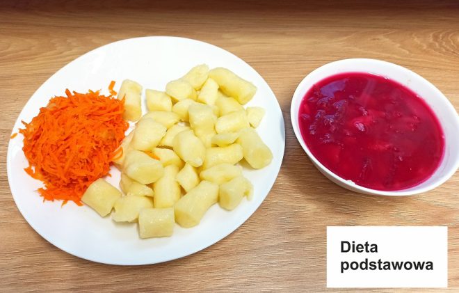 Na zdjęciu przedstawiono talerz z obiadem, obok którego umieszczono karteczkę z napisem: „Dieta podstawowa”. Na talerzu znajdują się kluski kładzione oraz porcja tartej marchewki w formie surówki. Obok na drugim talerzu podano barszcz czerwony z kawałkami buraków