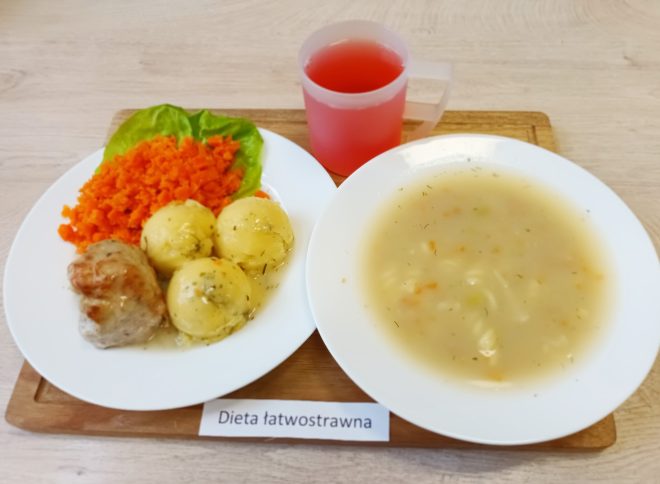 Na drewnianej desce przygotowano zestaw obiadowy z widoczną karteczką z napisem: „Dieta łatwostrawna”. Na jednym talerzu znajdują się trzy ziemniaki polane delikatnym sosem, marchewka na ciepło podana w formie drobnych kawałków oraz kawałek duszonego mięsa, ułożone na liściu sałaty. Na drugim talerzu podano klarowną zupę z kawałkami warzyw i dodatkiem koperku. Obok zestawu stoi kubek z czerwonym napojem