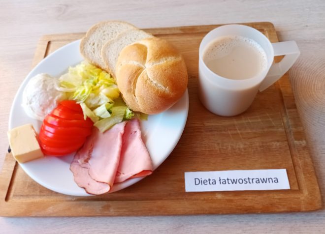 Na drewnianej desce znajduje się talerz z posiłkiem oznaczonym jako "Dieta łatwostrawna". Na białym talerzu umieszczono: świeże pieczywo (bułkę i dwie kromki chleba pszennego), dwa plastry wędliny, porcję startego lodowego sałaty, plasterek pomidora, kulkę twarogu i mały kawałek masła. Obok talerza stoi biały kubek z napojem mlecznym