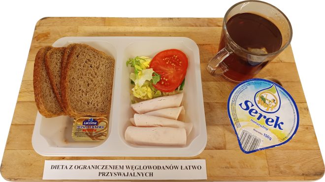 Zdjęcie przedstawia zestaw śniadaniowy z dietą ograniczającą węglowodany łatwo przyswajalne, serwowany na drewnianej desce. W plastikowym pojemniku znajduje się kilka kromek pełnoziarnistego chleba, plasterki chudej wędliny, sałata z plasterkiem pomidora oraz miniaturowa porcja masła. Obok zestawu leży kubeczek z serkiem naturalnym oraz szklanka z czarną herbatą. Na desce znajduje się kartka z napisem: 'Dieta z ograniczeniem węglowodanów łatwo przyswajalnych'. Zestaw wygląda zdrowo i jest przygotowany z uwzględnieniem potrzeb osób na specjalnej diecie