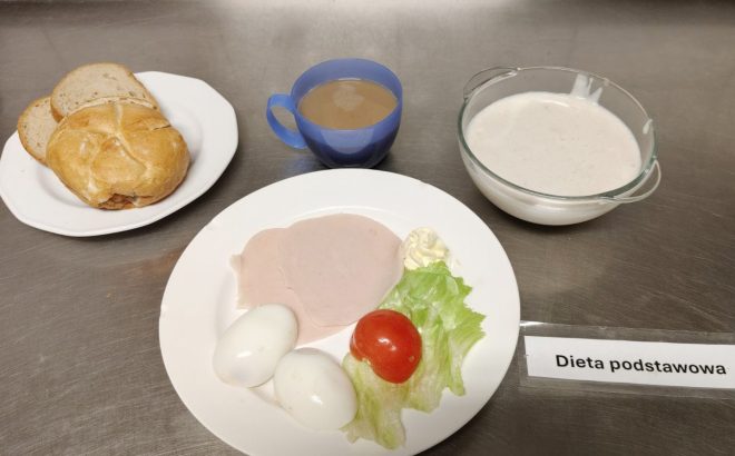 Na talerzu znajdują się dwie kromki pieczywa oraz bułka kajzerka. Obok na drugim talerzu podano dwa jajka na twardo, liść sałaty, plaster pomidora, dwie porcje wędliny drobiowej i niewielką porcję masła. W szklanym naczyniu zupa mleczna, a w niebieskim kubku napój, prawdopodobnie kawa z mlekiem. Danie oznaczone jest etykietą "Dieta podstawowa" i podane na metalowym blacie