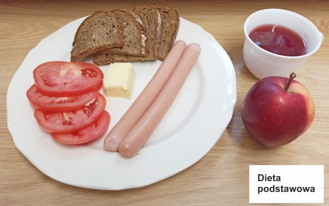 Na zdjęciu widoczny jest zestaw śniadaniowy oznaczony jako „Dieta podstawowa”. Na białym talerzu znajdują się trzy kromki chleba razowego, kawałek masła, plasterki świeżego pomidora oraz dwie cienkie parówki. Po prawej stronie talerza stoi kubek z czerwonym napojem, prawdopodobnie herbatą owocową, a obok niego leży czerwone jabłko