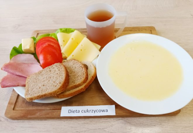 Na drewnianej desce podano zestaw śniadaniowy, obok którego widoczna jest karteczka z napisem: „Dieta cukrzycowa”. Na talerzu znajduje się kilka plasterków sera żółtego, dwa plasterki wędliny, świeże plasterki pomidora oraz mała kostka masła na liściu sałaty. Obok umieszczono dwie kromki chleba pełnoziarnistego. Na desce stoi również kubek z gorącą herbatą oraz talerz zupy mlecznej o jasnym, kremowym kolorze