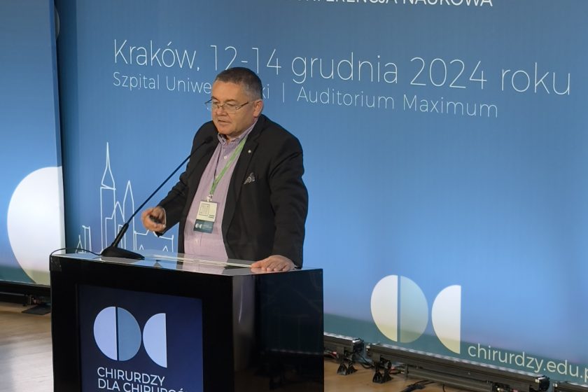 Starszy mężczyzna w garniturze i koszuli stoi za podium i wygłasza wykład podczas konferencji „Chirurdzy dla Chirurgów 2024” w Krakowie. W tle widoczny jest niebieski baner z nazwą wydarzenia, datą „12-14 grudnia 2024” i lokalizacją „Szpital Uniwersytecki | Auditorium Maximum”. Podium ma nowoczesny design z logo wydarzenia i grafiką przedstawiającą zarys architektury Krakowa