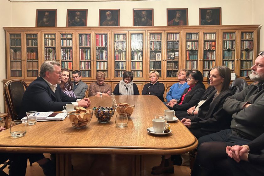 Na zdjęciu widać grupę osób siedzących przy dużym drewnianym stole w bibliotecznej sali konferencyjnej. Mężczyzna po lewej stronie prowadzi rozmowę, a pozostali uczestnicy słuchają z powagą. W tle stoją regały pełne książek za przeszklonymi drzwiami, a nad nimi zawieszone są obrazy przedstawiające postacie historyczne. Na stole znajdują się filiżanki z kawą, miski z ciasteczkami i cukierkami oraz karafki z wodą