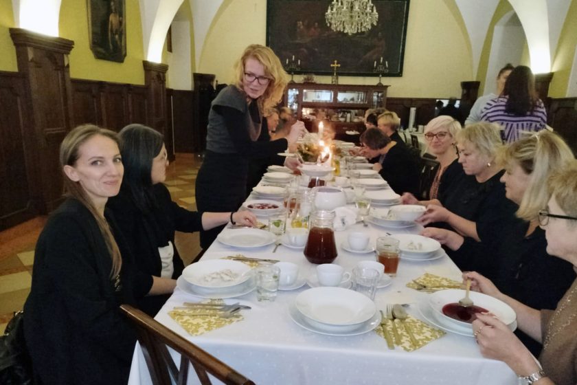 Zdjęcie przedstawia uroczysty posiłek przy długim, odświętnie nakrytym stole w sali jadalnej z łukowatym sklepieniem. Przy stole siedzą głównie kobiety, ubrane elegancko, zajmujące się jedzeniem i rozmowami. W tle widać ciemne, drewniane boazerie, świeczniki oraz stojący zegar. Na stole znajdują się talerze z barszczem, wazony z napojami i dekoracje świąteczne