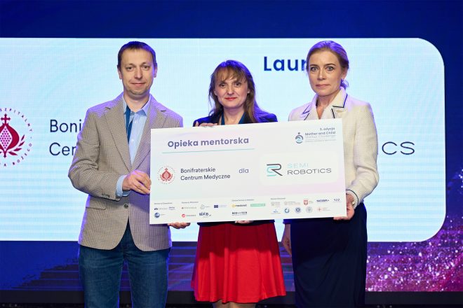 Troje laureatów na scenie podczas uroczystej gali pozuje do zdjęcia, trzymając dużą tablicę informacyjną. Tablica zawiera informacje o mentoringu między Bonifraterskim Centrum Medycznym a „SEMI Robotics”. Po lewej stronie stoi mężczyzna w jasnej marynarce, pośrodku kobieta w czerwonej spódnicy i ciemnej marynarce, a po prawej kobieta w jasnym żakiecie. W tle widoczne są logotypy wydarzenia i partnerów projektu