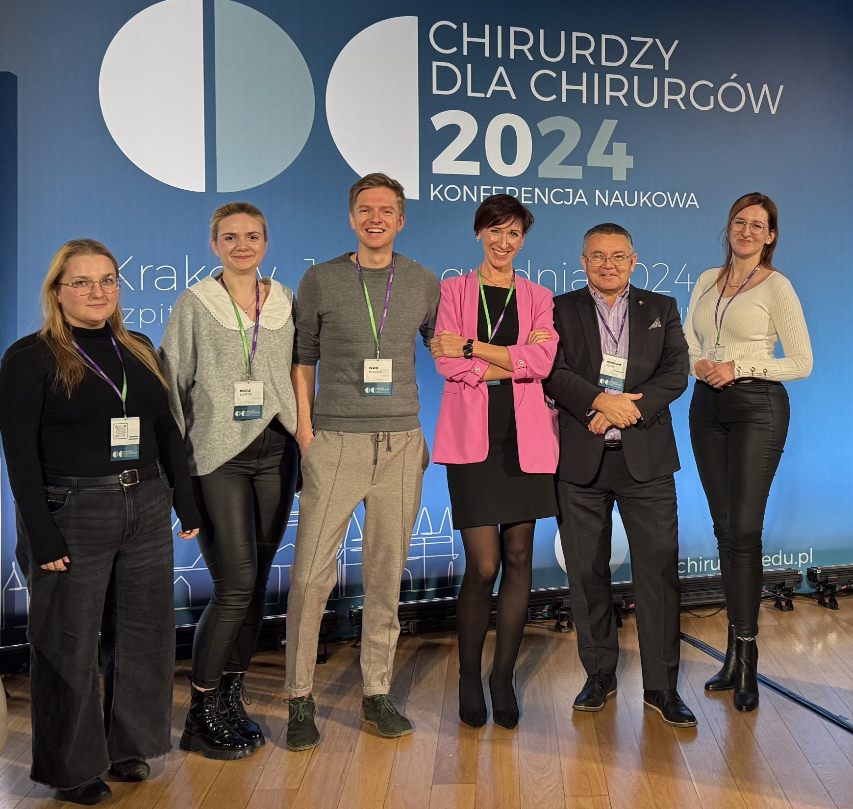 Grupa sześciu uczestników konferencji naukowej „Chirurdzy dla Chirurgów 2024” pozuje na scenie na tle dużego, niebieskiego baneru z logo wydarzenia i datą „12-14 grudnia 2024 roku”. Widać nowoczesną scenografię z oświetleniem, roślinnością i podium po prawej stronie. Uczestnicy stoją w swobodnych, profesjonalnych strojach, uśmiechając się do obiektywu. W tle po lewej widoczny jest pusty fotel i stolik z wodą