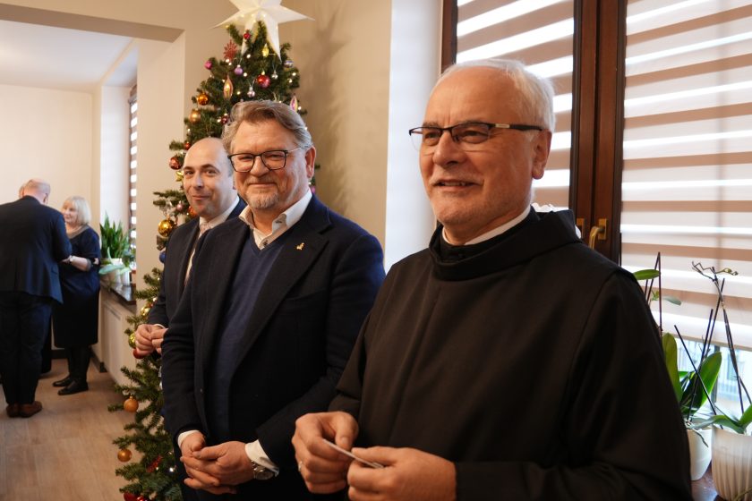 Grupa osób uczestnicząca w spotkaniu opłatkowym. W tle widoczna jest udekorowana choinka ze świątecznymi ozdobami. W centrum zdjęcia Ojciec Prowincjał uśmiecha się do aparatu, stojąc obok dwóch elegancko ubranych mężczyzn