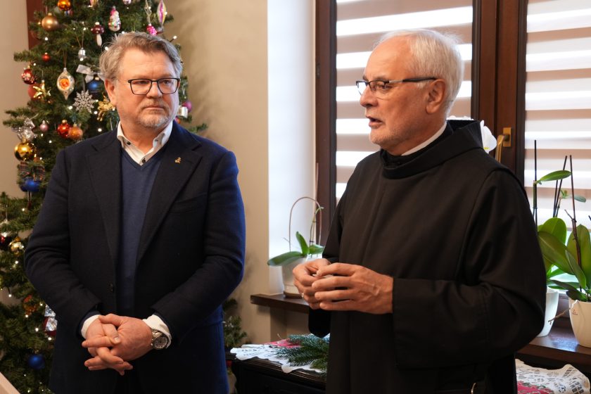 Ojciec Prowincjał wygłasza przemówienie, gestykulując rękoma. Obok niego stoi mężczyzna w eleganckim granatowym garniturze, z uwagą słuchając. W tle znajduje się udekorowana choinka i okno z roletami