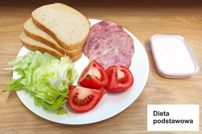 Na drewnianym blacie znajduje się zestaw kolacyjny oznaczony jako „Dieta podstawowa”. Na talerzu znajdują się trzy kromki chleba, liście sałaty lodowej, kawałki pomidora oraz plasterki wędliny drobiowej z dodatkami. Obok ustawiono pojemniczek z białym sosem lub dipem