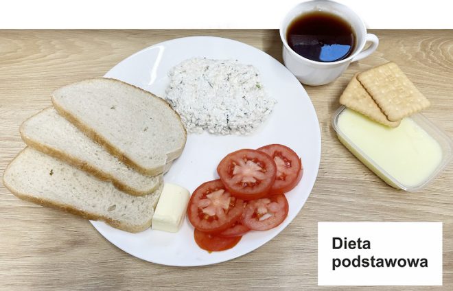 Zdjęcie przedstawia zestaw kolacyjny oznaczony jako „dieta podstawowa”. Na talerzu znajdują się trzy kromki chleba, kawałek masła, plasterki świeżego pomidora oraz porcja twarożku z dodatkiem szczypiorku. W tle ustawiono filiżankę z herbatą, a obok pojemnik z budyniem waniliowym i dwoma herbatnikami