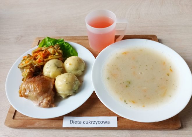 Na drewnianej podstawce ustawiony zestaw obiadowy oznaczony jako "Dieta cukrzycowa". Na białym talerzu znajdują się trzy ziemniaki w sosie koperkowym, kawałek pieczonego kurczaka oraz surówka z duszonej fasolki szparagowej z marchewką. Obok znajduje się miseczka zupy jarzynowej o kremowej konsystencji z widocznymi kawałkami warzyw. Całość uzupełnia przezroczysty kubek z różowym napojem