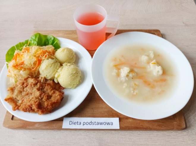 Na jednym talerzu znajduje się panierowany kotlet schabowy, trzy kulki ziemniaków puree oraz surówka z kapusty kiszonej i marchewki ułożona na liściu sałaty. Obok, w białym głębokim talerzu, jest zupa kalafiorowa z kawałkami kalafiora i marchewki w jasnym bulionie. Po lewej stronie deski umieszczono prostokątną karteczkę z napisem „Dieta podstawowa”. Do posiłku dołączony jest przezroczysty plastikowy kubek z czerwonym kompotem