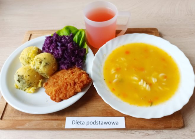 Na drewnianej tacy podano zestaw obiadowy oznaczony jako "Dieta podstawowa". W jego skład wchodzą: talerz zupy jarzynowej o jasnopomarańczowym kolorze z dodatkiem makaronu świderki, podany na białym głębokim talerzu. Obok znajduje się talerz z daniem głównym – trzy ugotowane ziemniaki posypane koperkiem, panierowany kotlet schabowy o złocistej barwie, porcja gotowanej czerwonej kapusty oraz liść sałaty. Na tacy stoi także przezroczysty plastikowy kubek z różowym napojem