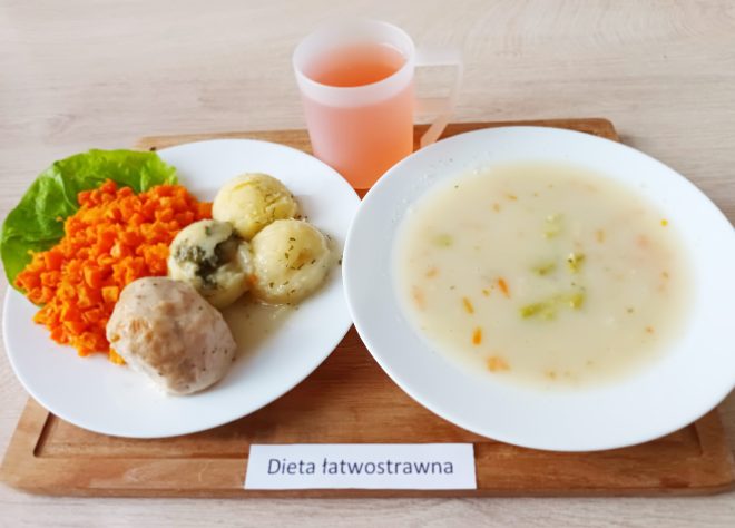 Na drewnianej tacce znajduje się zestaw diety łatwostrawnej. Po lewej stronie białego talerza znajduje się porcja duszonej marchewki, obok niej dwie małe kulki ziemniaków z ziołami oraz niewielki kawałek mięsa z sosem. Po prawej stronie umieszczony jest talerz z jasną, delikatną zupą warzywną z dodatkiem marchewki i brokułów. W tle przezroczysty kubek z różowym napojem. Na środku tacki widnieje podpis „Dieta łatwostrawna”