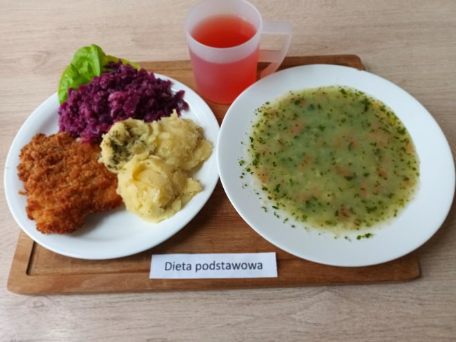 Na zdjęciu przedstawiono obiad w ramach diety podstawowej. Na pierwszym talerzu znajduje się panierowany kotlet schabowy, porcja puree ziemniaczanego z koperkiem, a także duszona czerwona kapusta z dodatkiem liścia sałaty. Na drugim talerzu podano klarowną zupę z zieleniną i kawałkami warzyw. Obok znajduje się przezroczysty kubek z różowym napojem. Na drewnianej podstawce pod talerzami leży biała kartka z napisem „Dieta podstawowa”