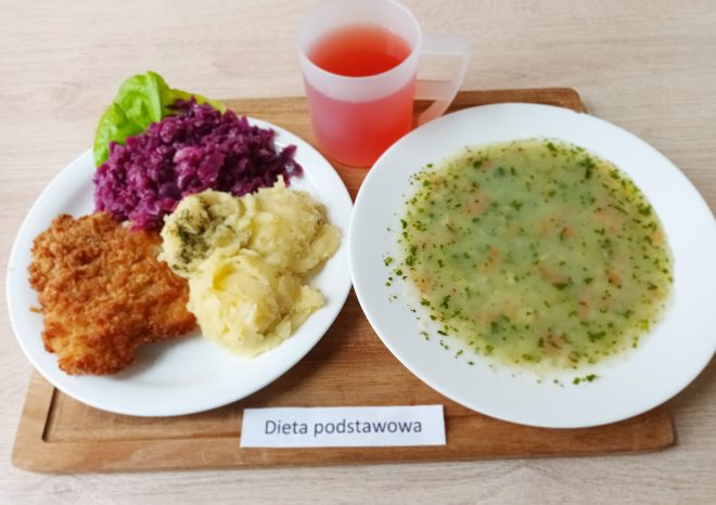 Na drewnianej tacy widoczne jest danie obiadowe oznaczone jako „Dieta podstawowa”. Na talerzu po lewej stronie znajduje się kotlet schabowy w złocistej panierce, porcja ziemniaków puree z odrobiną koperku oraz czerwona kapusta na ciepło. Na talerzu po prawej stronie znajduje się zielona zupa jarzynowa z pietruszką i kawałkami warzyw. Obok stoi przezroczysty kubek z czerwonym napojem, prawdopodobnie kompotem