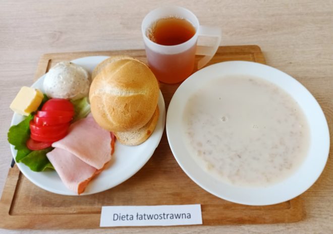 Na drewnianej tacy znajduje się zestaw śniadaniowy w ramach diety łatwostrawnej, co wskazuje umieszczona poniżej kartka z napisem: "Dieta łatwostrawna". Na białym talerzu znajduje się chrupiąca bułka, dwa plastry wędliny, kawałek masła, liść sałaty, trzy plasterki pomidora i porcja twarożku. Obok stoi przezroczysty kubek z ciepłą herbatą. Na osobnym talerzu podano kaszę mannę na mleku