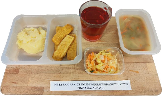 Na zdjęciu przedstawiono posiłek oznaczony jako „Dieta z ograniczeniem węglowodanów łatwo przyswajalnych”. W plastikowym pojemniku podano porcję puree ziemniaczanego oraz trzy panierowane paluszki rybne. Obok w osobnej przegródce znajduje się zupa jarzynowa z kawałkami warzyw, a w małym pojemniczku surówka z marchwi i kapusty. W tle stoi szklanka z czerwonym napojem. Wszystko ustawiono na drewnianej desce z nazwą diety