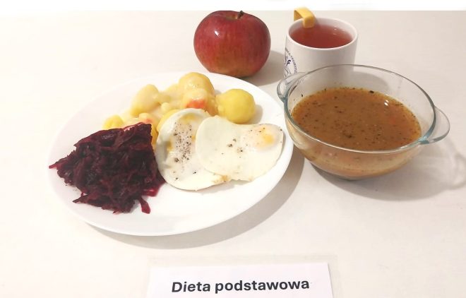 Na zdjęciu widać obiad oznaczony jako "Dieta podstawowa". Na białym talerzu znajdują się: dwie porcje jajek sadzonych posypanych pieprzem, gotowane ziemniaki z sosem i warzywami (marchewką i kalafiorem) oraz buraczki na ciepło w formie tartej. Obok talerza stoi szklana miska z zupą o ciemnobrązowym kolorze z widocznymi przyprawami i warzywami. Uzupełnieniem posiłku jest jabłko oraz biały kubek z herbatą, z plasterkiem cytryny na krawędzi