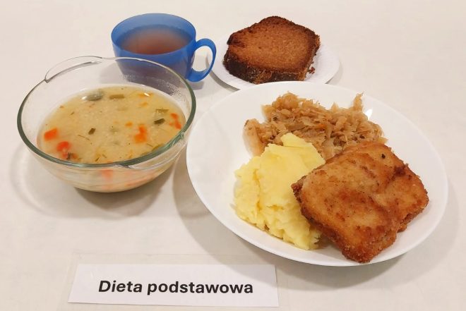 Na zdjęciu przedstawiono posiłek oznaczony jako „Dieta podstawowa”. Na białym talerzu znajdują się panierowana ryba, puree ziemniaczane oraz duszona kapusta. W przezroczystej miseczce podano zupę jarzynową z kawałkami warzyw. Obok ustawiono niebieski kubek z herbatą oraz kromkę ciemnego pieczywa