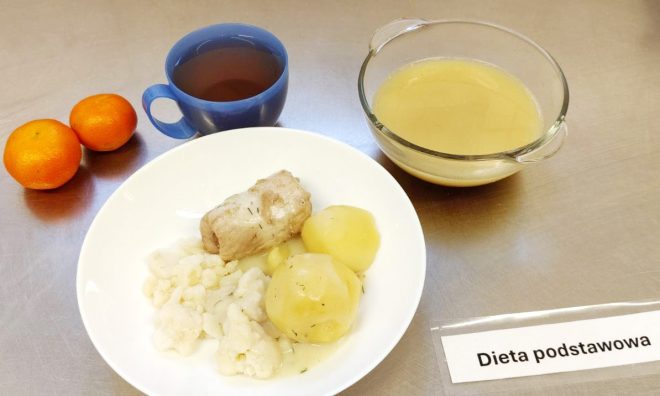 Zdjęcie przedstawia zestaw obiadowy oznaczony jako „dieta podstawowa”. Na talerzu znajdują się ziemniaki z koperkiem, różyczki kalafiora i kawałek mięsa gotowanego. Obok ustawiono miskę z przezroczystego szkła z bulionem, kubek z napojem (prawdopodobnie herbatą) oraz dwie mandarynki