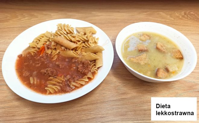 Na drewnianym blacie znajduje się zestaw obiadowy oznaczony jako „Dieta lekkostrawna”. Na talerzu po lewej stronie podano pełnoziarnisty makaron typu penne i świderki z sosem pomidorowym oraz kawałkami mięsa mielonego i warzyw. Obok, w misce, znajduje się zupa krem z dodatkiem grzanek