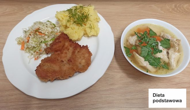 Przedstawiono obiad w ramach diety podstawowej. Na talerzu znajdują się ziemniaki puree posypane koperkiem, panierowany kotlet schabowy oraz surówka z białej kapusty z marchewką i koperkiem. Obok talerza stoi miseczka z rosołem ozdobionym natką pietruszki. Na dole zdjęcia widoczny jest napis „Dieta podstawowa”